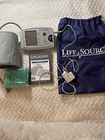 Photo showing blood pressure monitor display, cuff, instruction manual, pill organizer, and carrying bag laid out on a beige textured surface.