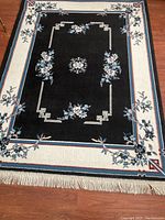 Full view of rectangular floral area rug with black center and cream border with blue and rose floral accents and fringed edges.