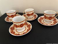12 art deco teacups and saucers with distinctive orange and brown geometric patterns, gold-trimmed handles, arranged on black surface.
