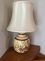 Full view of a ceramic table lamp with a hand-painted floral base and beige fabric lampshade on a wooden surface against a beige wall.