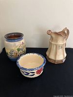Three ceramic items displayed together: a cityscape decorated vase, a folk art decorated bowl, and a beige floral ceramic jug.