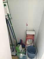 Photo shows various cleaning tools stored in corner including corn broom, multiple dustpans, blue bucket, fuzzy duster in box, and mop handles.