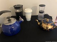 Blue stovetop kettle with black handle, black manual coffee grinder, white-cup coffee maker, and vintage green and gold Gibson & Sons teapot displayed on black surface.