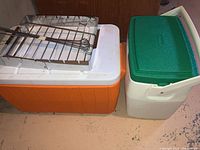 Two plastic coolers, one orange and white and one white and green, with metal BBQ skewers and tray placed on top.