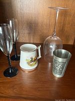 Photo showing two crystal champagne flutes with black twisted stems, one large clear wine glass, English Prince William ceramic mug with gold trim, and a pewter tumbler cup