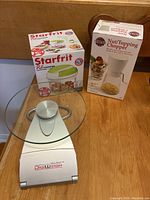 Photo showing Digiweigh digital kitchen scale with glass platform in front, and boxed Starfrit Pull Chopper and Norpro Nut/Topping Chopper behind on a wooden surface.