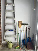 7-foot aluminum step ladder leaning against wall with gardening tools arranged beside it, including two brooms, shovels, weed diggers, and a green watering can on the floor.