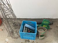 Photo showing two tall cylindrical wire tomato cages, a blue plastic milk crate, green handheld seed spreader labeled 'Scotts', and a grey plastic pot inside the crate with hose and nozzle items nearby