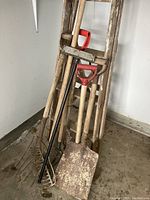 Full view of the gardening tools including ladder, shovels, pitchfork, and rake leaning against the wall in a garage corner.