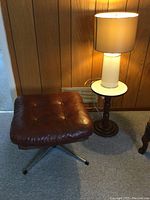 Brown leather-like cushioned swivel foot stool with button tufting and metal base, placed beside wood side table and lamp
