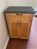 Front view of kitchen cart showing wooden build, black speckled top, one drawer above double doors with handles, and caster wheels at bottom.