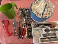 The kitchen items and dinnerware laid out on a pink table covering including utensils, a green milk jug, disposable plates, disposable cutlery, and kitchen tools such as a garlic press and wine openers.