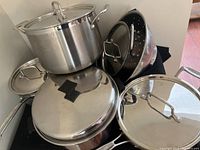 Photo showing a large stainless steel stock pot with lid, stack of pots and pans with lids, and a black perforated steamer or colander insert.