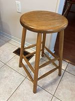 Side view of the stool showing wooden round seat, legs, and support bars on tiled floor.