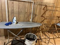 View of the ironing board with gray cover, Proctor Silex iron with blue soleplate on top, laundry basket in front, and metal clothing racks behind all against wood-paneled wall.
