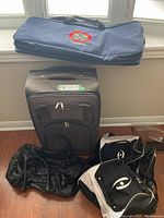 Photo showing all four items: two black duffel bags, one black wheeled suitcase, and one navy blue squash racquet bag, arranged on wooden floor against window