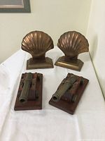 Photo showing four bookends: two brass clamshell designs and two bronze cannon on wooden bases.