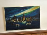 Frontal photo of the cityscape painting clearly showing the artwork, metal frame, and signature, with visible hues of blue, green, yellow, and city lights reflecting on water.