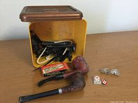 Two wooden tobacco pipes in front of a yellow container filled with pipe parts, along with a pair of gold toned playing card themed cufflinks and two vintage enamel pins.