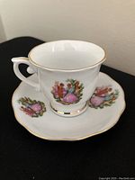 White porcelain teacup and saucer with colorful Victorian courting couple design, scalloped edges, and gold trim on a black surface.