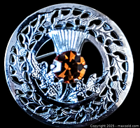 Close-up photo of round silver-tone Mizpah kilt pin with central citrine gemstone amid detailed thistle leaf and flower carvings.