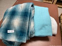 Three folded blankets stacked showing blue and white plaid Eddie Bauer fleece throw on top, light blue textured blanket, and white knitted blanket beneath