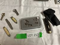 Collection of small folding knives and pocket cutters with cream and black handles, metal nail clipper, and a black pouch on a white surface