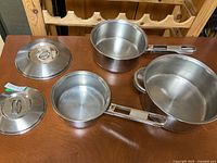 Three stainless steel pots with matching lids on wooden table from an angled top view showing sizes and handles.