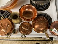 Multiple copper kitchen pieces including decorative embossed pans, small copper bowl, brass vase, and small brass ladle arranged on stove.