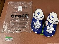 Pair of blue and white men's Crocs with Toronto Maple Leafs logos and their packaging bag on a brown flat surface.