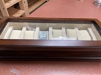Front angled view of wooden Bombay watch box on reddish floor, showing glass top and white padded cushions inside, holding one watch.