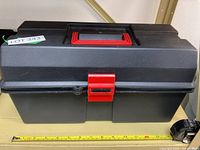 Closed black plastic tool box with red latch and handle shown on a measuring tape to indicate size.