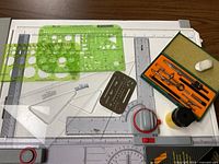 Full view of drafting board with various drawing tools including a green stencil, triangular rulers, metal curve stencil, drafting tool case, ink pot, and eraser.