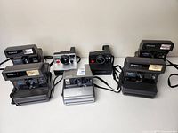 Overview of all seven Polaroid cameras on a white surface, showing variety of models and colors including black and silver bodies.