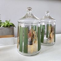 Two clear glass apothecary jars decorated with alternating vertical green stripes and gold leaf designs, each with a round lid and knob handle.