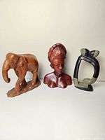 Photo showing three distinct carved sculptures on a white background: a wooden elephant, a female wooden bust, and an abstract black sculpture with grey accents.