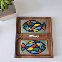 Top-down photo of Mid-Century Modern wooden tray with two ceramic tiles depicting colorful abstract fish and silver-tone center handle.