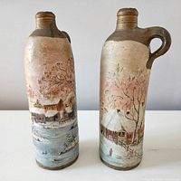 Front side of two handpainted tall stoneware jugs showing winter cottage and snowy landscape scene
