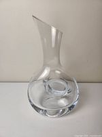 Clear glass decanter with wide rounded body and angled open neck against plain background.