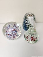 Three hand blown glass paperweights displayed on a white surface, showing colorful internal designs and clear glass body.