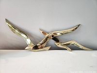 Three images showing a large brass wall hanging sculpture of three abstract birds in flight, polished reflective gold tone, minimalist design.