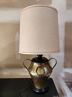 Front view of the large hammered brass base lamp with beige lampshade, showing the decorative brass ring and handles.