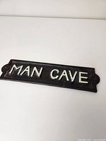 Full view of rectangular cast iron 'MAN CAVE' sign with raised white letters on black background.
