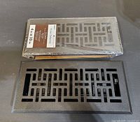 Two metal floor vent covers, one new in packaging, one unpackaged, shown flat on surface with geometric pattern and black finish visible