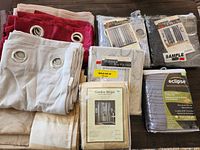 New drapery panels in multiple colors and styles stacked on a wooden floor with metal grommet tops visible on some panels.