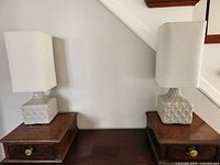 Pair of ceramic table lamps shown side by side on wooden tables, against a wall with stairs in the background.