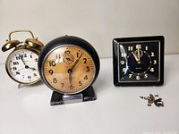Front view of three vintage alarm clocks including brass bell, black round clock, and black square clock with keys on the right side