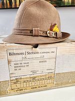 The vintage grey felt Fedora hat with decorative feather detail and stitched band, positioned on top of its original box with label showing name and address of Mr. Young the Tailor.