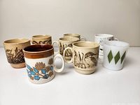 Seven vintage mugs with varied designs including ceramic stoneware with nature motifs and Fire King milk glass mugs with green diamonds and text
