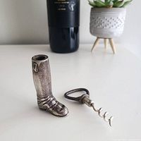 Silver riding boot shaped container and detached corkscrew open on white surface with bottle in background.
